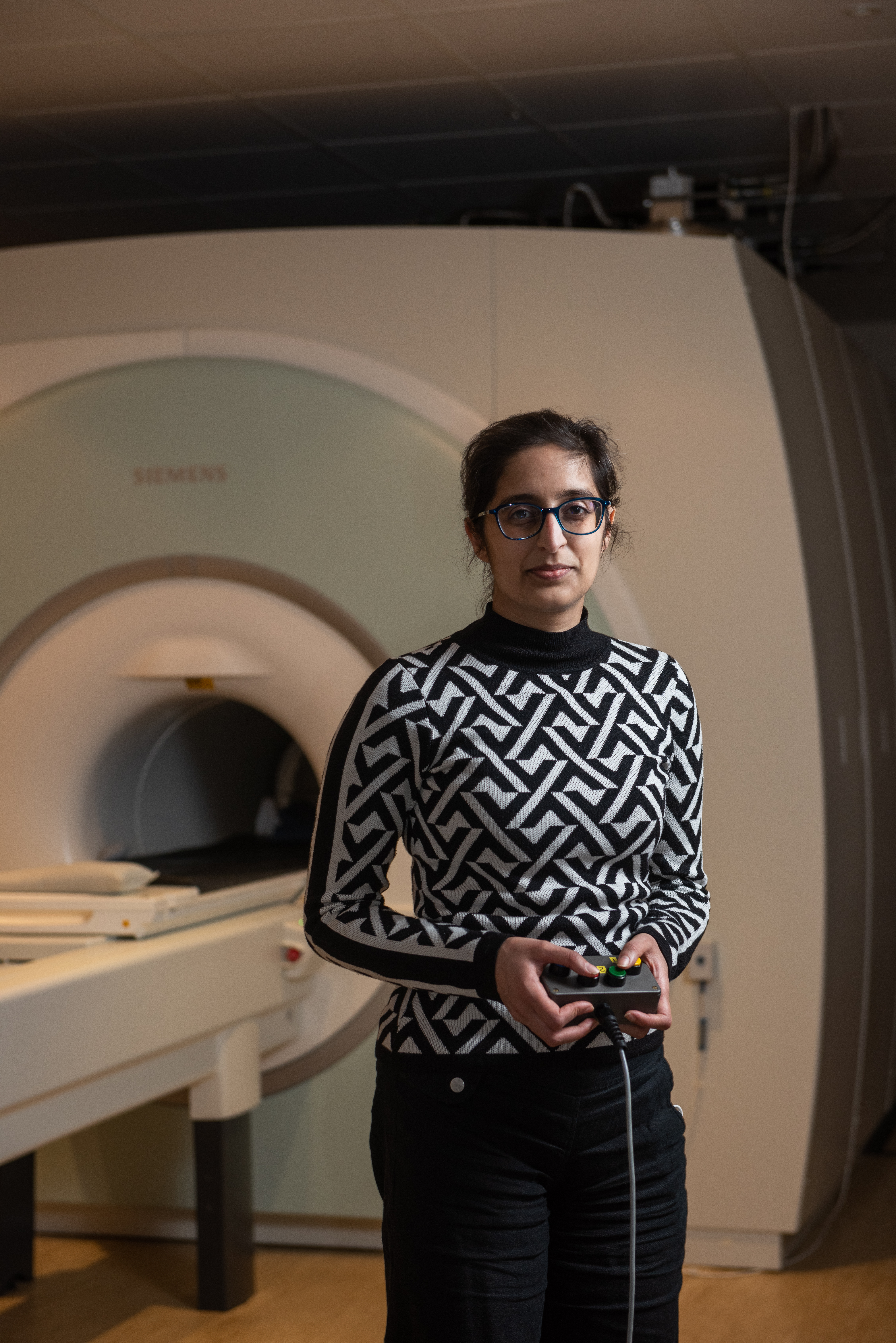 Dr Jasleen Jolly, stood in front of an MRI machine. She is wearing a black and white diamond pattern jumper and black glasses. She has dark hair and is holding the machine controls.