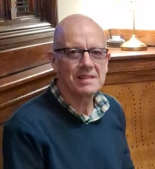 Macular Society volunteer Chris Norris, sat down wearing glasses and a shirt underneath a dark blue jumper.