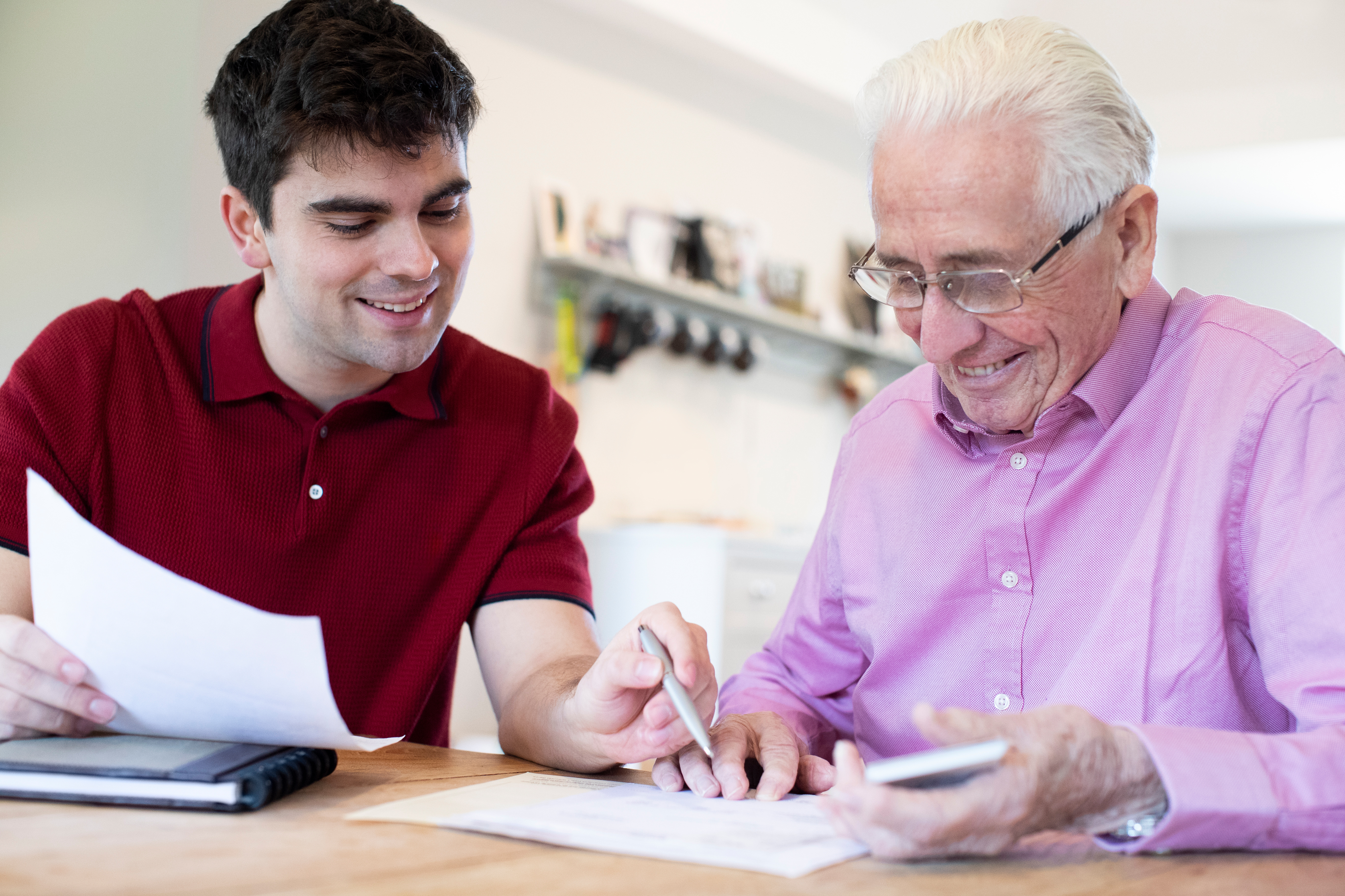One young white man and one elderly white man sat at a table looking at paperwork together, smiling 