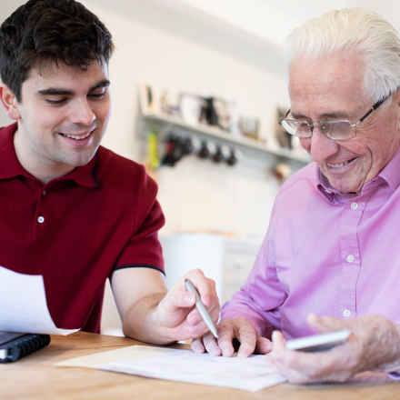 One young white man and one elderly white man sat at a table looking at paperwork together, smiling 