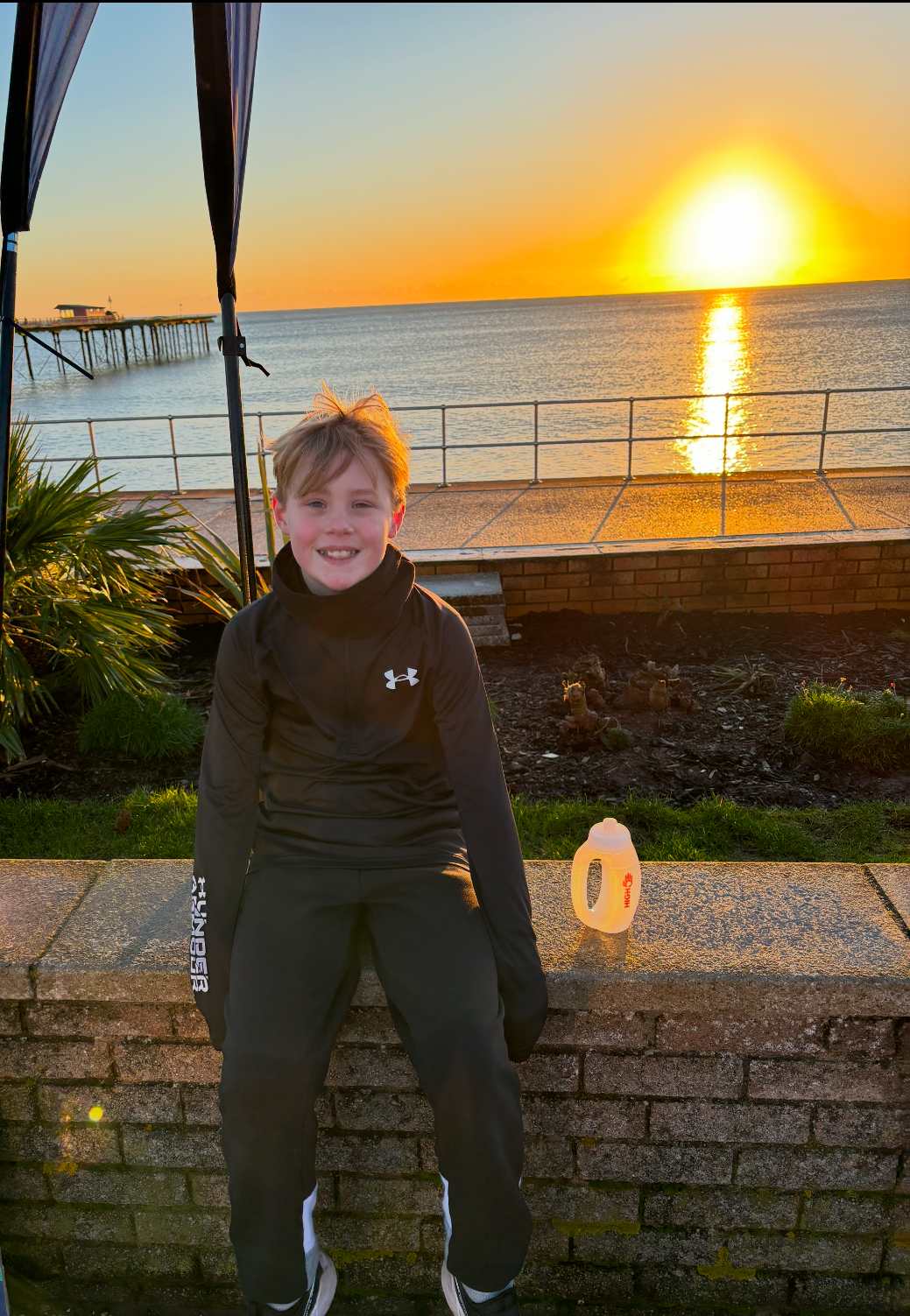 Picture shows young 10 year old Archie sitting on a wall at the coast with sunset in the background 