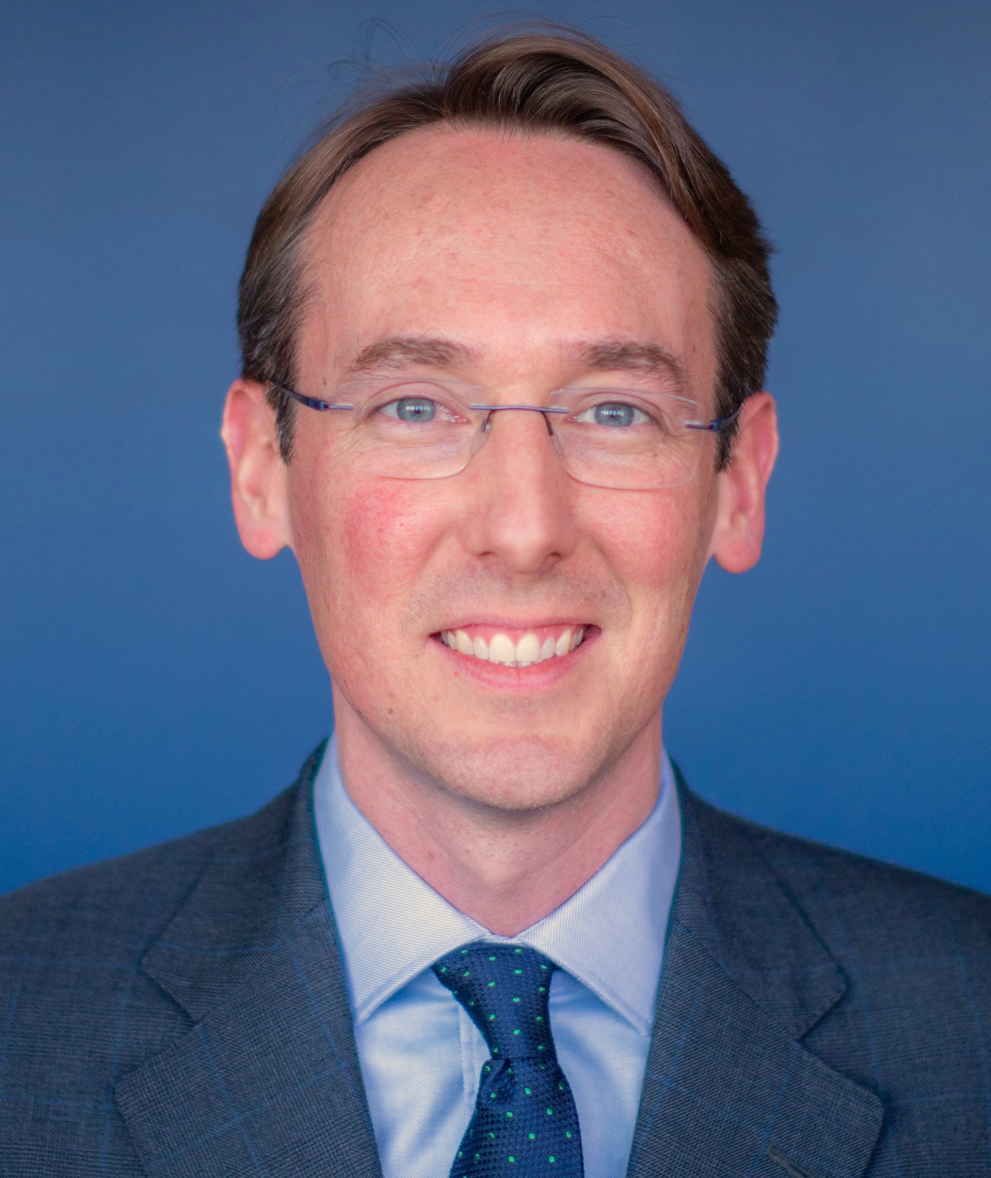 Headshot of Dr Keenan, a white man smiling at the camera wearing glasses and shirt, tie and suit. he is sitting against a blue background