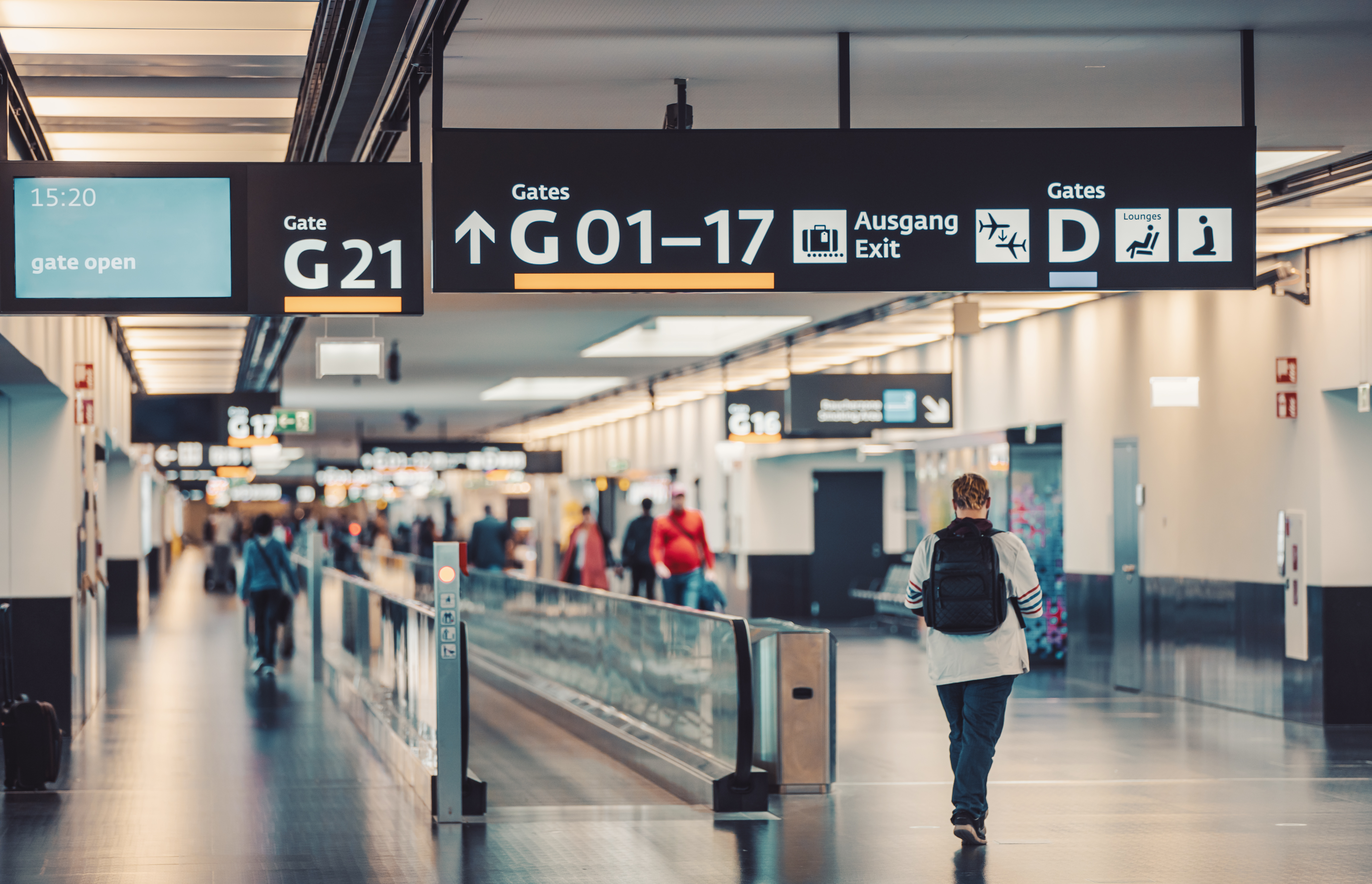 Departures at an airport. The middle of the image is a walking conveyer belt and a person walking towards it. Above are the gate numbers.
