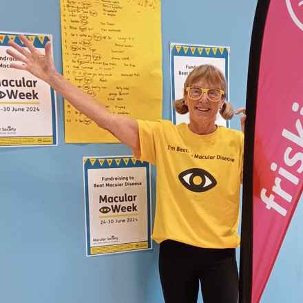 Fitness leader Rosey with her arm aloft wearing a Macular Society fundraising t-shirt. Macular Week posters are in the background