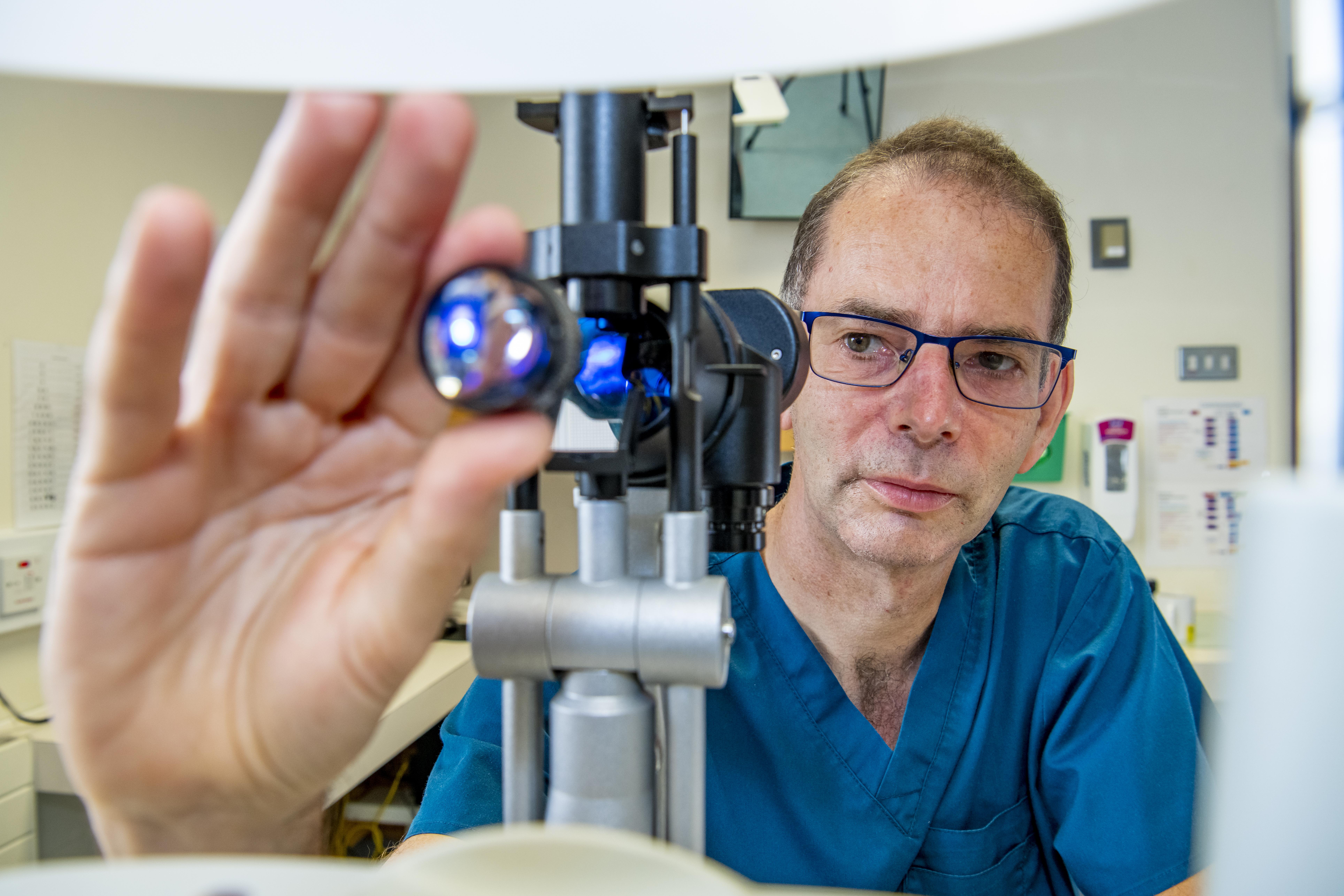 Andrew Lotery using optometrist equipment. Andrew is wearing blue scrubs and glasses and has dark short hair.