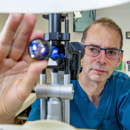 Andrew Lotery using optometrist equipment. Andrew is wearing blue scrubs and glasses and has dark short hair.