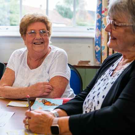 Two woman sit around a table, arms crossed while smiling