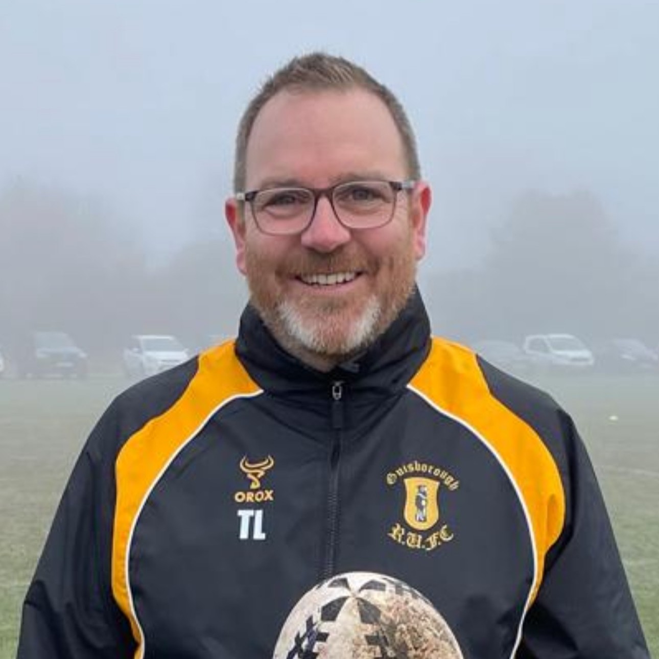 Trevor has short light brown hair and glasses, smiling down the camera outside on a foggy day. He is wearing his rugby club's dark coloured jacket with orange stripes coming down from each shoulder. The jacket includes the Guisborough Rugby Club crest and his initials. The top half of a rugby ball can also be seen which Trevor is holding.
