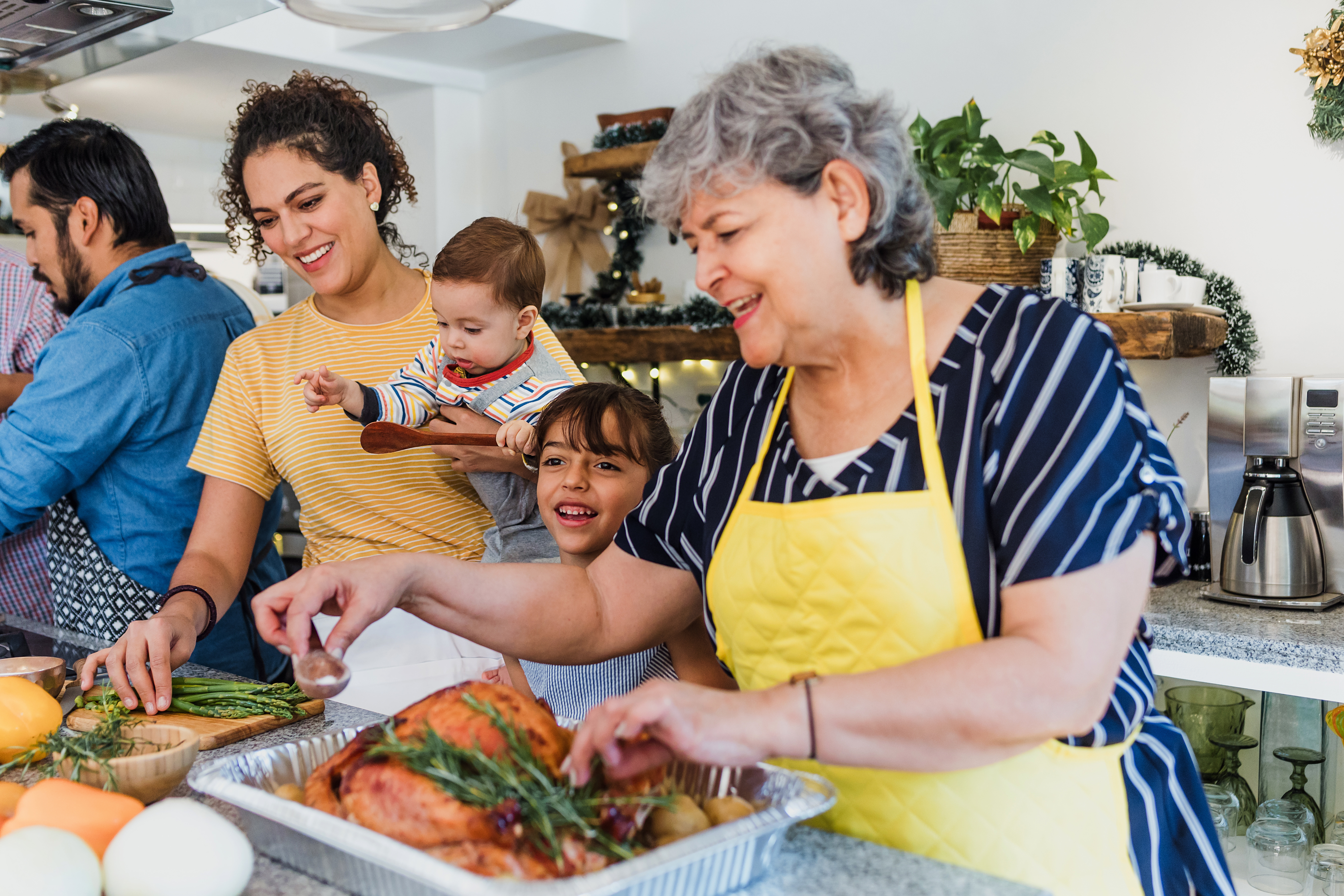 Family Christmas scene. Older woman in apron preparing a cooked turkey, smiling, surrounded by family, including a young girl and a baby being held by his mother who is helping to prepare food 