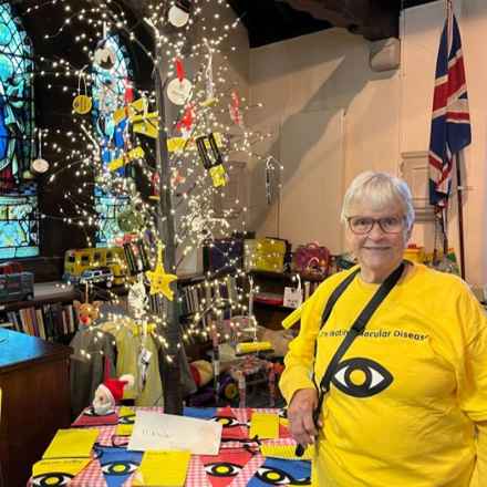 Jenny wearing her yellow Macular Society fundraising t-shirt stands next to the Macular Society-inspired Christmas tree, as part of her local church's Christmas Tree festival. The tree is a small, thin branched tree decorated with festive ornaments including a Santa, Rudoplh and a yellow star as well as items from the Macular Society online store, including the visual impairment badge and lined paper.