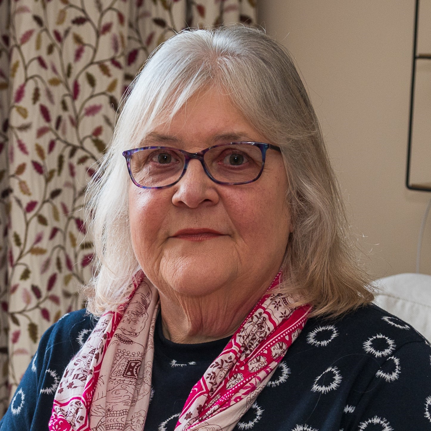 Headshot of Lynne. She is indoors, wearing a navy top with white circular patterns and a red scarf.