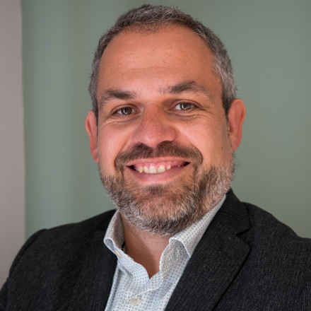 A head and shoulders shot of Ed Holloway, a white man with dark/grey hair smiling to camera, wearing a dark grey suit jacket and white open necked shirt