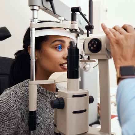A woman with black hair tied back and a grey jumper has a laser shone in her eye during an eye exam