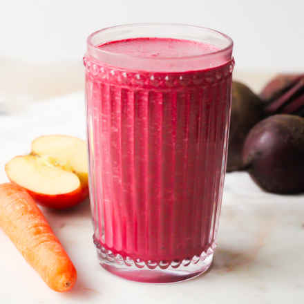 A red/purple coloured smoothie pictured in a clear glass with a carrot and apple chopped in half in the background 
