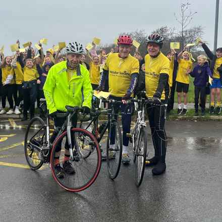 Gerard McCarten, middle, wearing his yellow Macular Society fundraising t-shirt and red helmet poses with his bike, flanked by his friends Mike Cowley and Ged Brear. Behind, Jericho Primary School children cheer and wave.