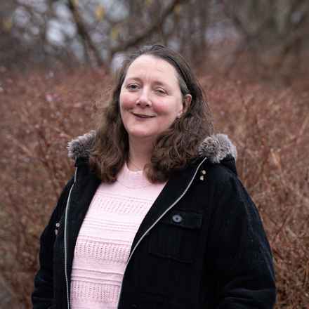 Kate who has PIC, smiling as she stands looking at the camera. She is outside, in front of a bare hedge. Kate has dark shoulder-length hair, and is wearing a pink jumper and hooded jacket.