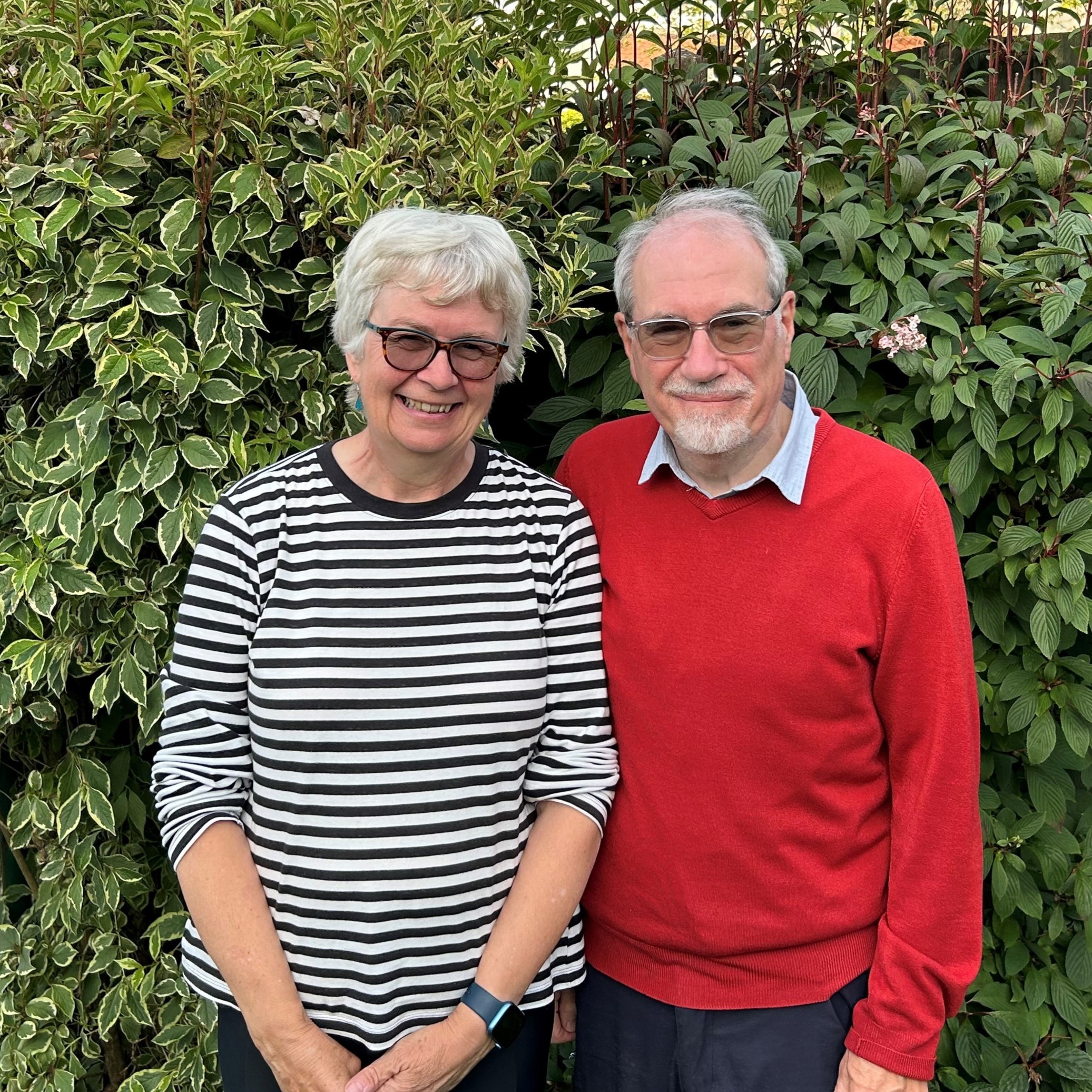 Jane, left, wearing glasses and a horizontal striped jumper, standing next to her husband Ed. He is also wearing glasses and a red jumper.