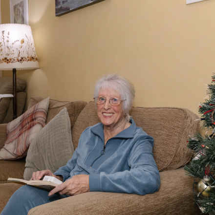 Margaret Hughes smiling in living room, reading on sofa