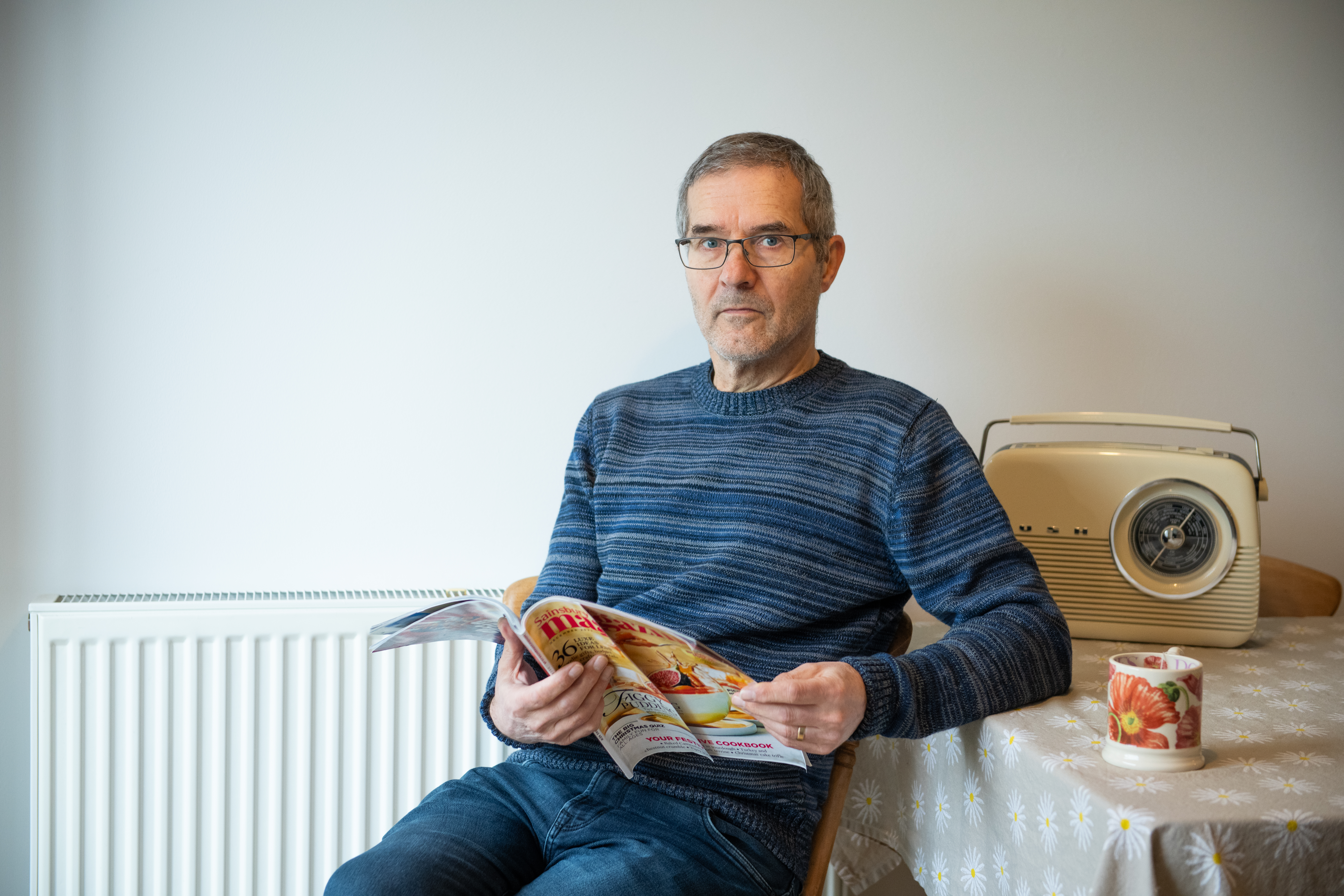 Simon staring at camera, while holding a magazine. A radio and cup of tea sit on the table