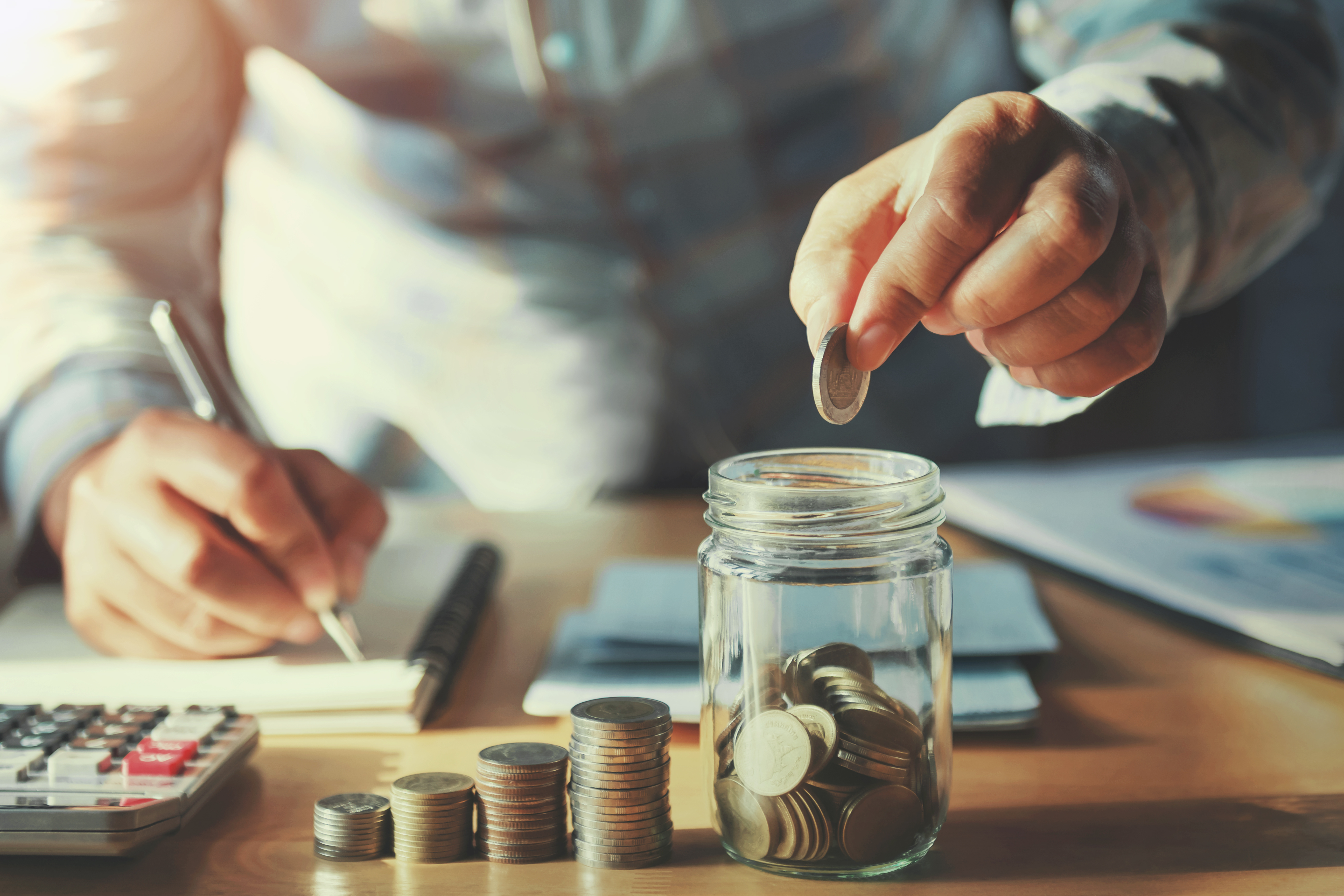 A man counting his money, while writing down calculations. There are four piles of coins, each growing in size, next to a jar of coins.