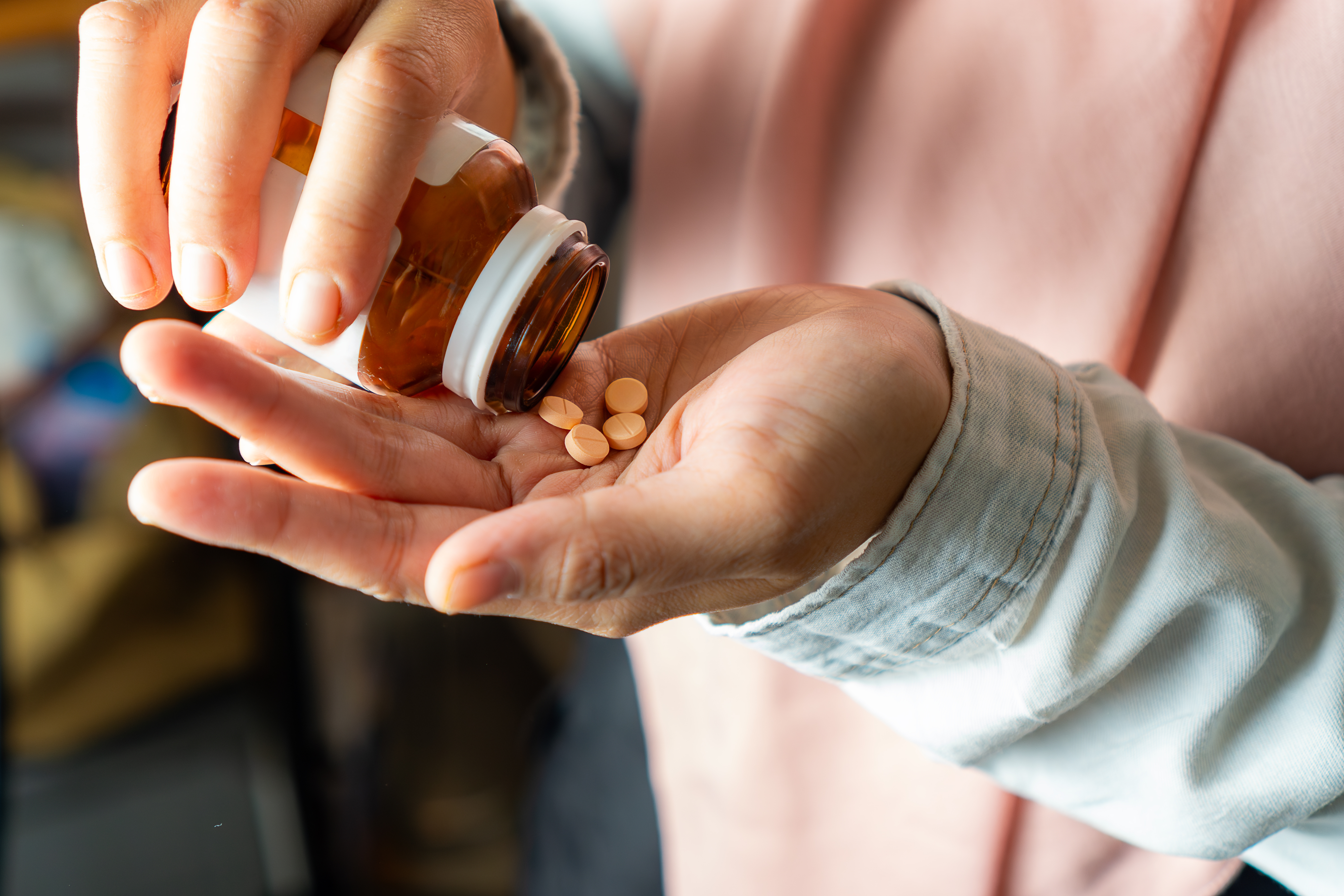 One hand holding a pill bottle tipping the pills out into an open hand, which holds two pills. 